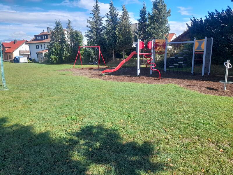 Ein Spielplatz mit einer roten Rutsche, Schaukeln und Klettergerüsten befindet sich neben einer Reihe von Bäumen und einigen Wohnhäusern.