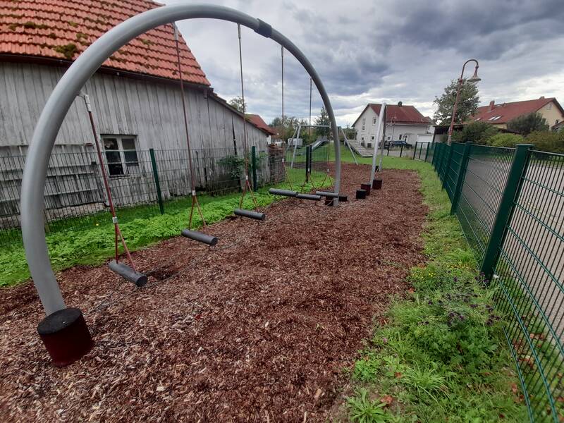 Ein Spielplatz mit Schaukelgestängen ohne Schaukelsitze ist von einem Gitterzaun und einem schrägen Holzdachgebäude umgeben.