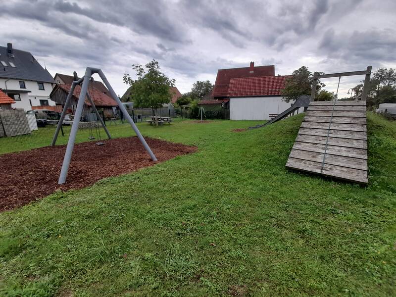Ein Spielplatz mit einer Schaukel links und einer Rutsche auf einem kleinen Hügel rechts, umgeben von Häusern und Bäumen bei bewölktem Himmel.