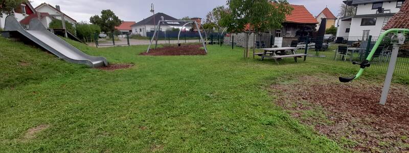 Eine grüne Parklandschaft mit Rutsche, Schaukel und Picknicktisch, umgeben von Häusern.