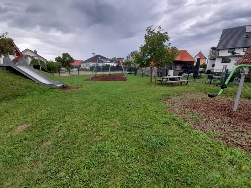 Eine grüne Parklandschaft mit Rutsche, Schaukel und Picknicktisch, umgeben von Häusern.