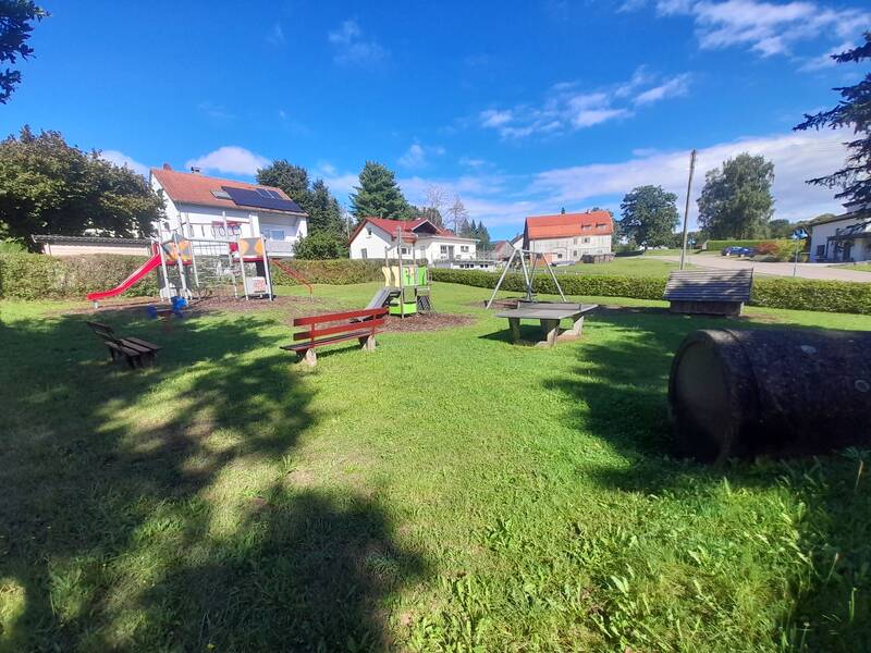 Ein kleiner Spielplatz auf einer Wiese mit Rutsche, Schaukel, Sitzbänken und einem Holzrohr, umgeben von Wohnhäusern im Hintergrund.