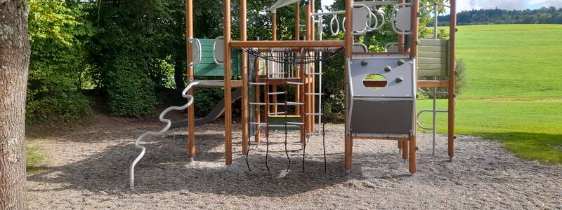 Ein moderner Spielplatz mit Klettergerüst steht auf einem kiesbedeckten Untergrund, umgeben von grünen Bäumen und Wiesen.