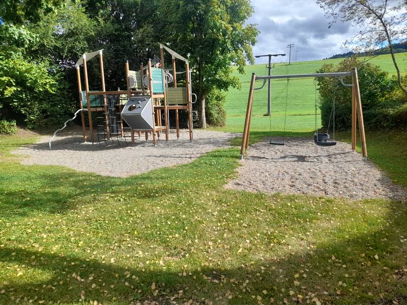 Ein Spielplatz mit einem Klettergerüst und zwei Schaukeln, umgeben von grüner Landschaft und Bäumen.