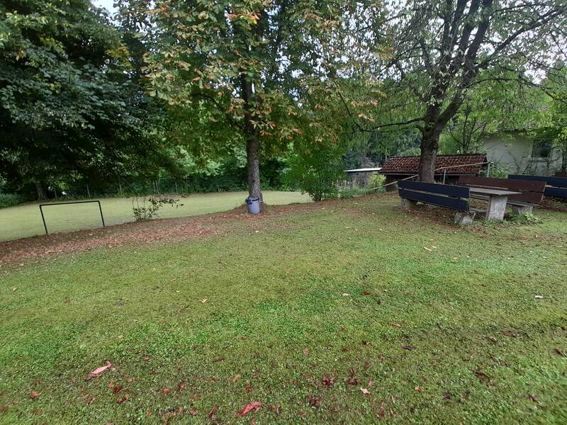 Ein grüner Park mit Bäumen, einem Fußballtor und zwei Bänken steht unter wolkigem Himmel.