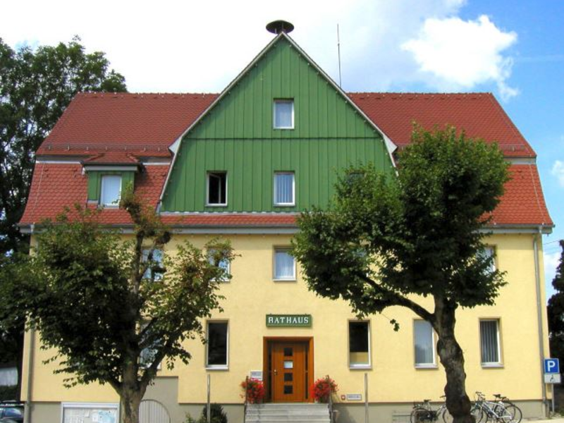 Das Bild zeigt ein zweistöckiges, gelbes Rathaus mit rotem Dach und grünem Giebel, umgeben von Bäumen und einem Fahrradständer.