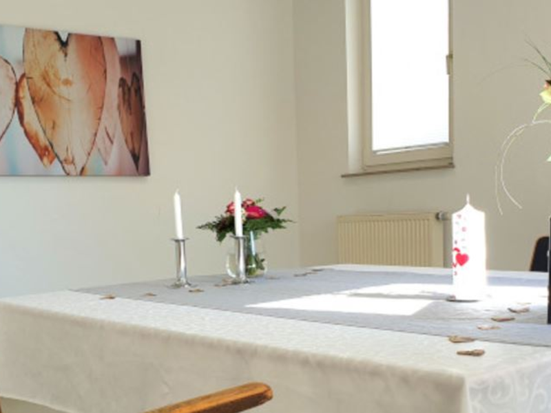 Ein helles, schlicht eingerichtetes Zimmer mit einem gedeckten Tisch, auf dem Kerzen und ein Blumenstrauß stehen, und einem Wandbild mit Herzen.