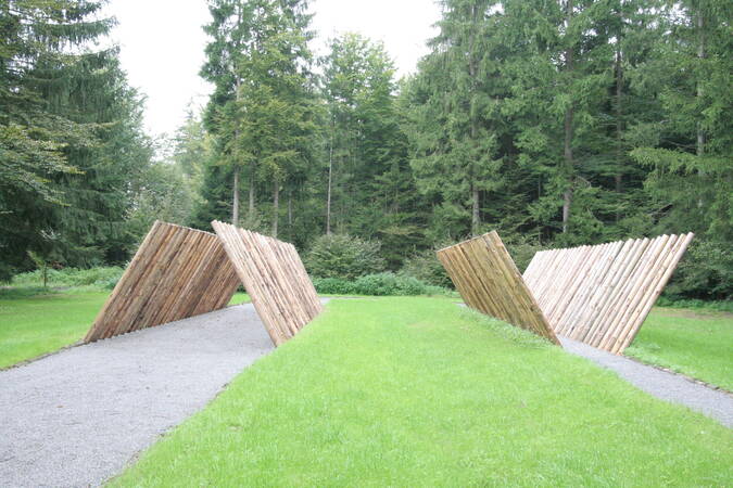 Zwei diagonale Holzkonstruktionen flankieren einen geschotterten Weg in einem grünen Waldgebiet.