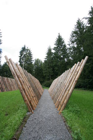 Ein schmaler Kiesweg führt durch eine Reihe schräg aufgestellter Holzstämme, umgeben von grünem Gras und hohen Bäumen im Hintergrund.