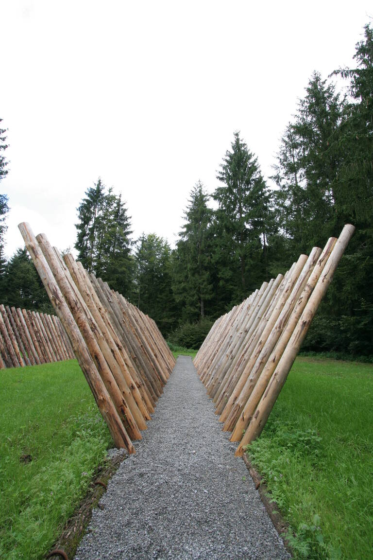 Ein schmaler Kiesweg führt durch eine Reihe schräg aufgestellter Holzstämme, umgeben von grünem Gras und hohen Bäumen im Hintergrund.