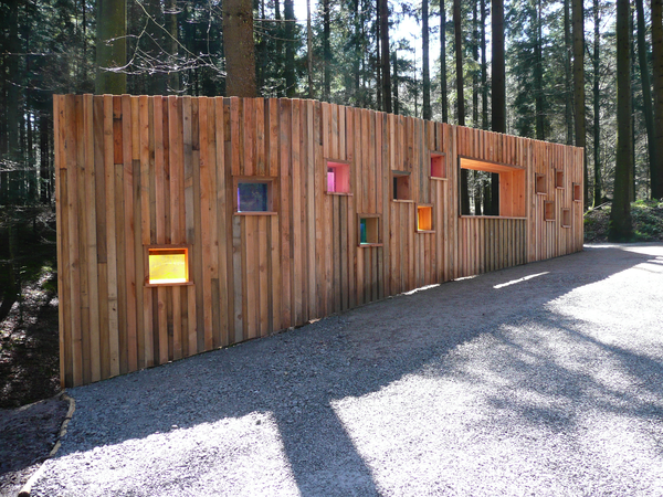 Eine hölzerne Wand mit mehreren kleinen, bunten Fenstern steht auf einem kiesigen Weg in einem bewaldeten Gebiet.