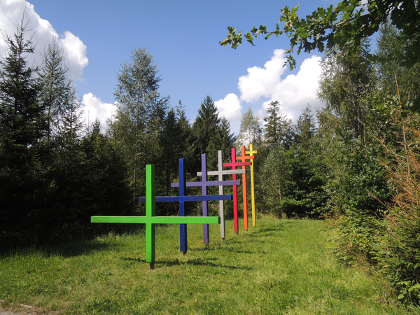 Mehrere farbige Holzkreuze stehen auf einer grünen Wiese, umgeben von Bäumen und unter einem blauen Himmel mit Wolken.